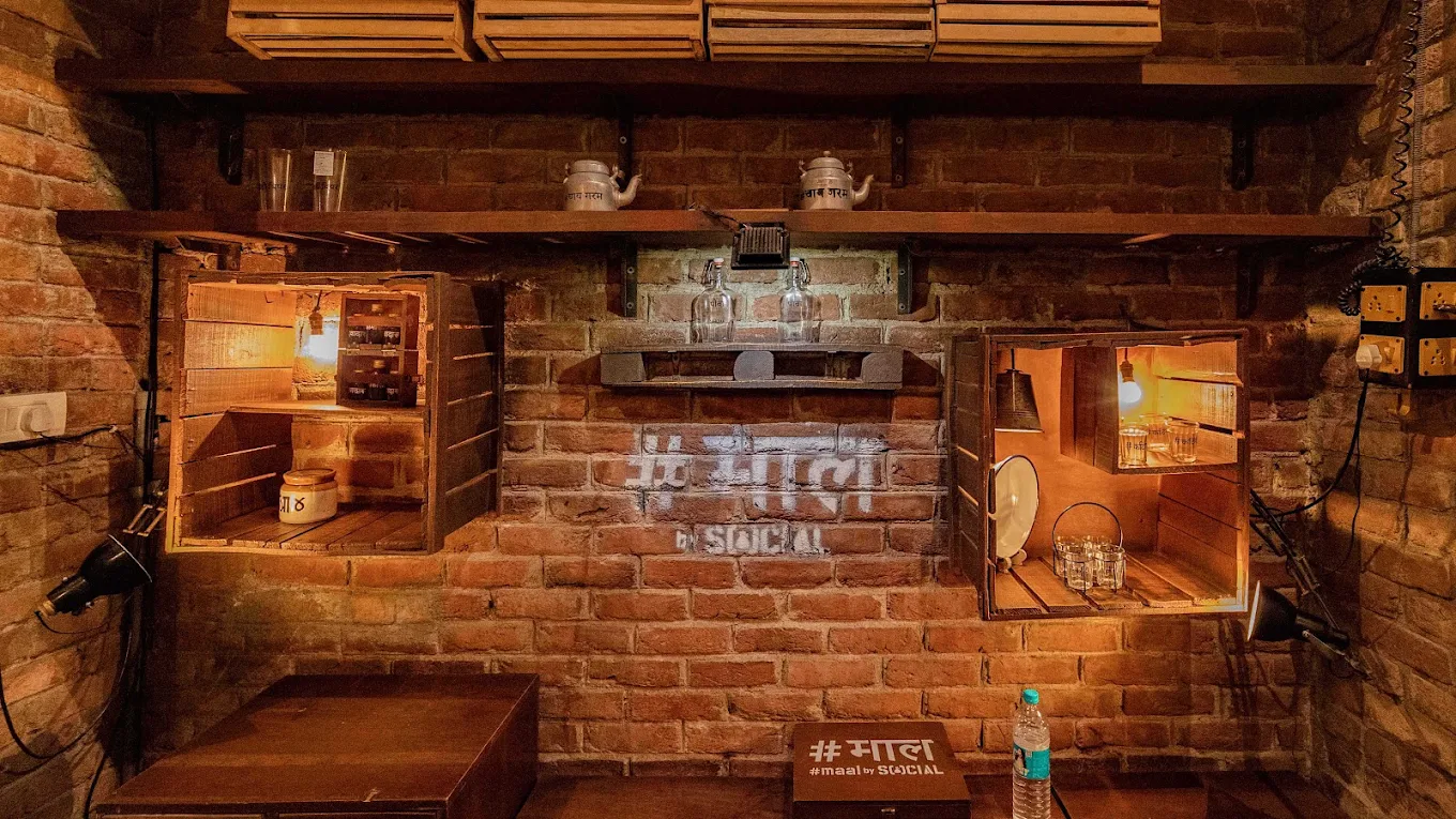 Rustic exposed-brick wall with warm-lit wooden crate shelves, brass teapots and glassware at Social, one of the cozy night pubs in Delhi