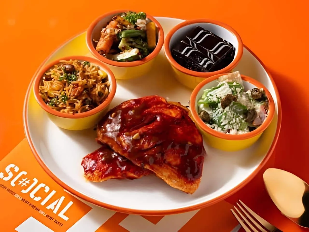 Plate of glazed chicken with four small bowls of rice, grilled vegetables, salad, and a chocolate brownie on an orange Social menu.